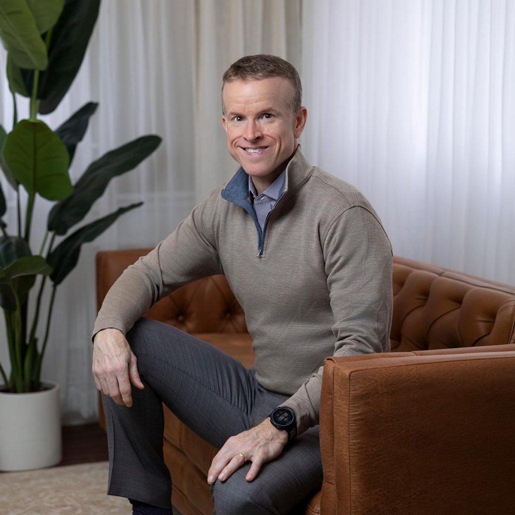 Dr. Kevin Hill seated in a leather chair in a professional setting, wearing a gray quarter-zip sweater and dress shirt, smiling warmly at the camera.